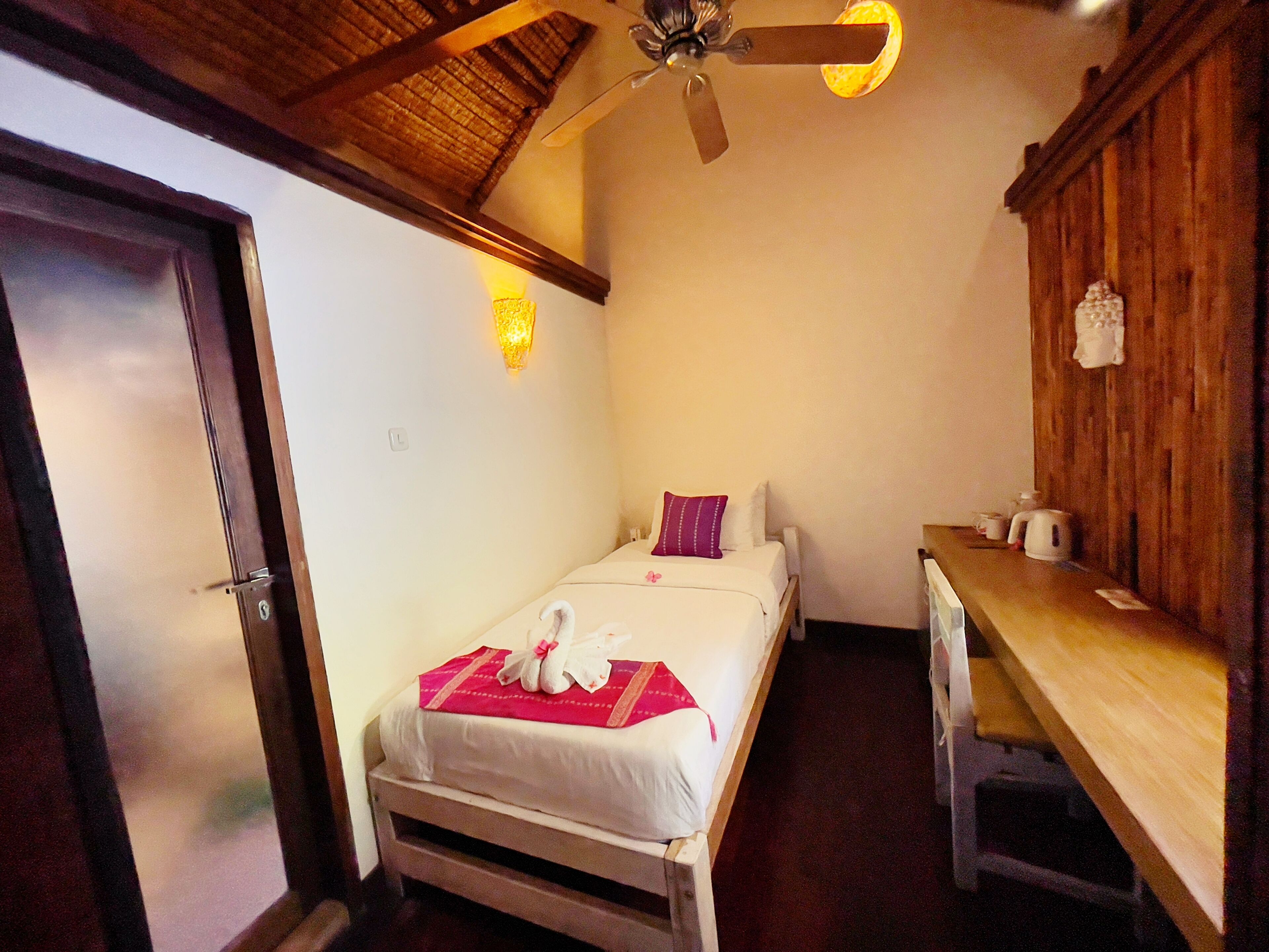 A small, cozy bedroom with a single bed, a wooden desk, a ceiling fan, and a wall-mounted light fixture.