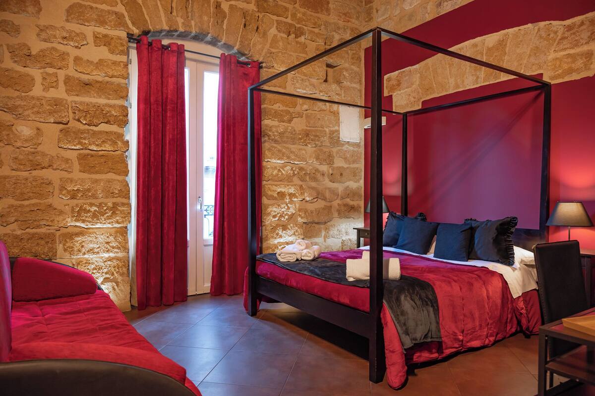 Una cama con dosel frente a una pared de piedra, con cortinas y cubrecama rojos.