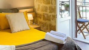 A bedroom with a stone wall, a bed with a yellow bedspread, a wooden headboard, a nightstand with a lamp, and a balcony with a chair. - B&B Carella (Palermo)