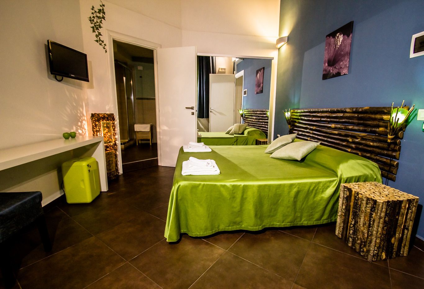 A modern hotel room with a green bed, a flat-screen TV, a wooden headboard, and a small wooden stool.