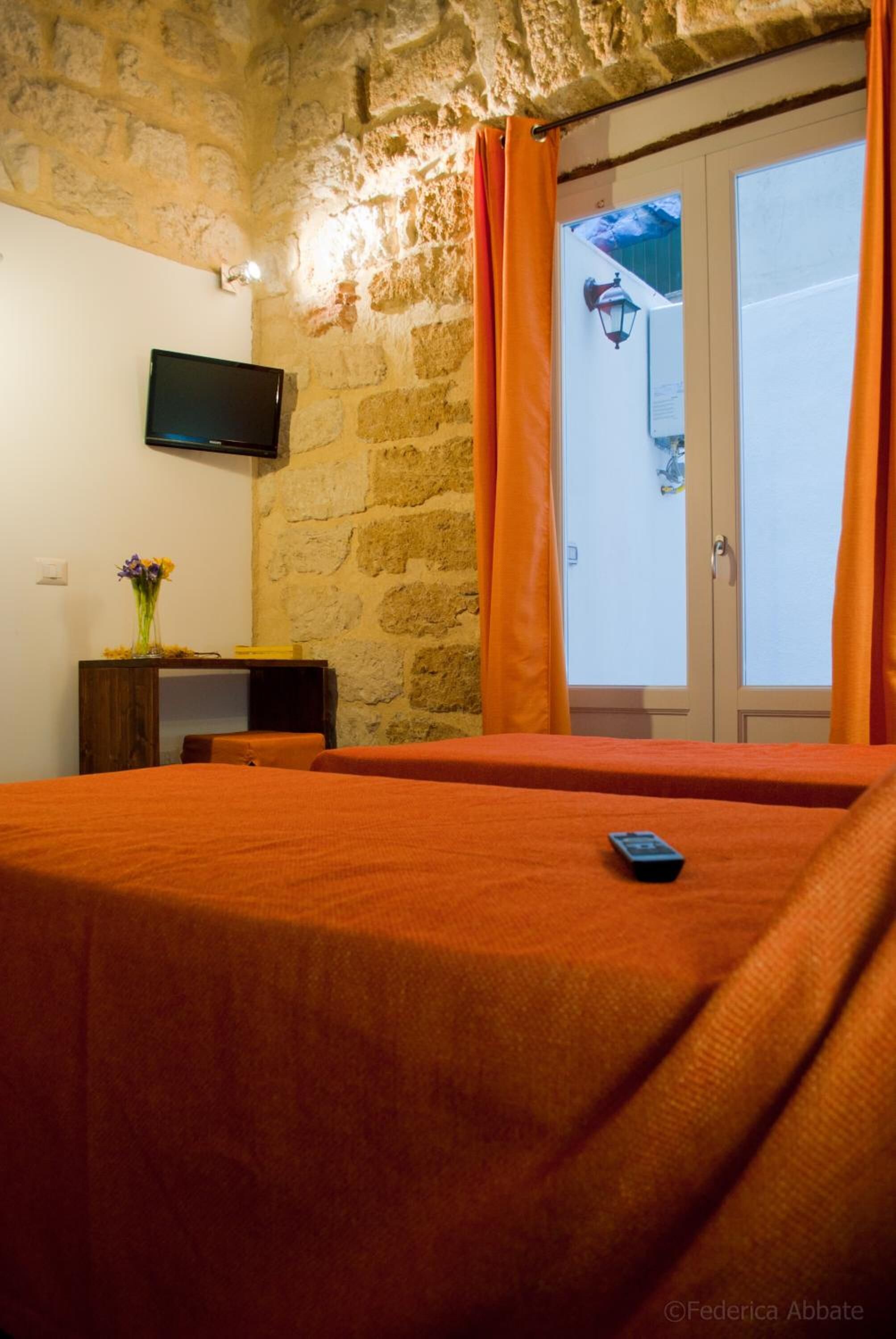 A room with a stone wall, a flat-screen TV mounted above a wooden table with a vase of flowers, and a bed with orange bedding.