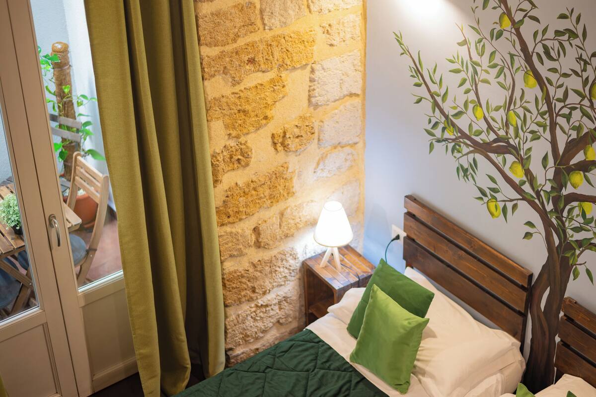 Una habitación con cama, cabecera de madera, mesita de noche con lámpara, pared de piedra y ventana con cortinas.