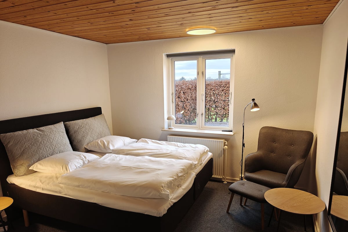 A bedroom with a bed, a chair, a small table, and a window with a view of trees.