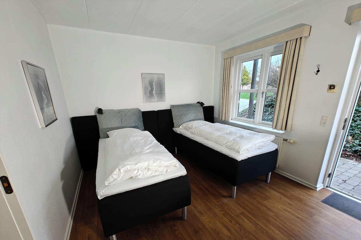 A room with two single beds, a window with curtains, a radiator, and a door leading outside.