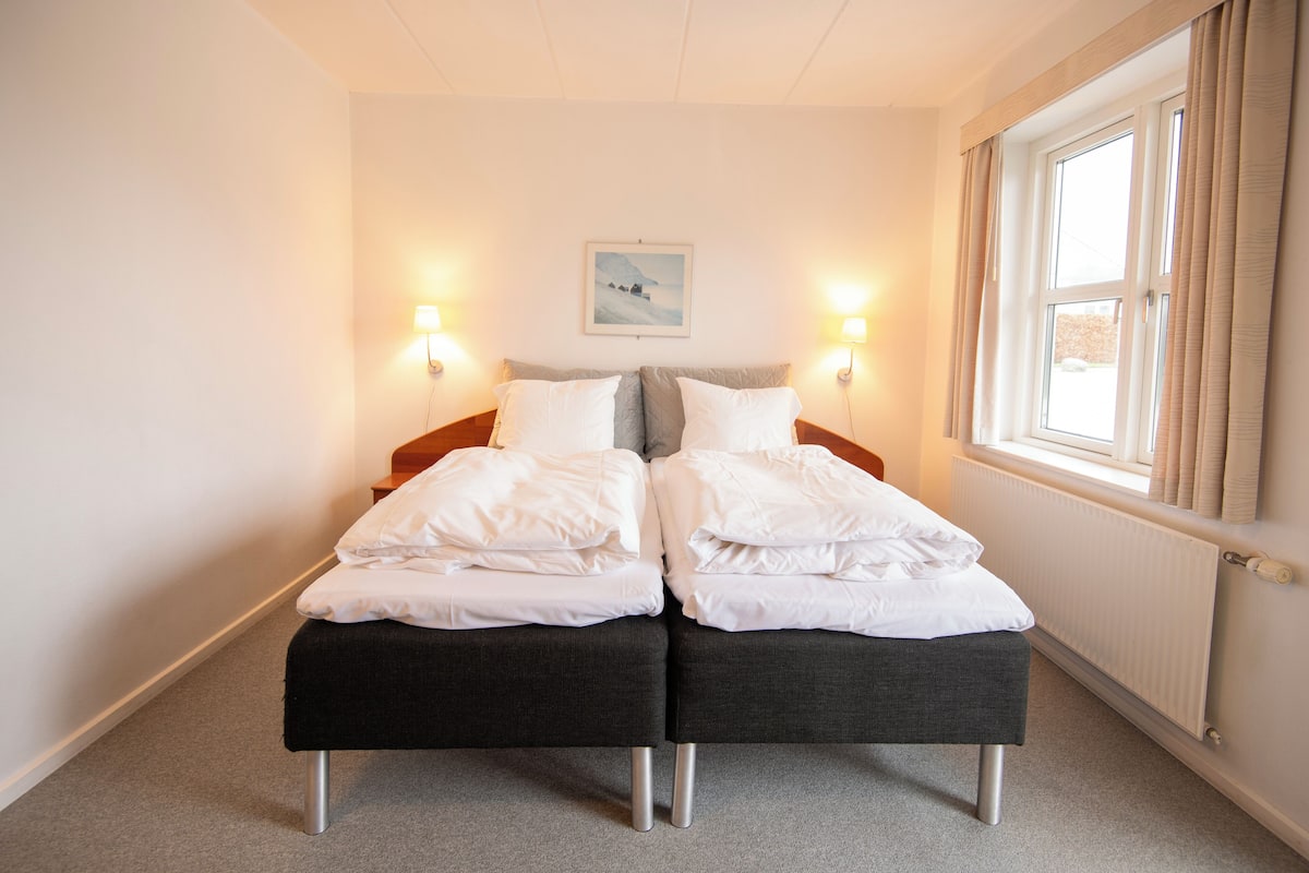 A double bed with white bedding, two bedside lamps, a framed picture on the wall, and a window with curtains.