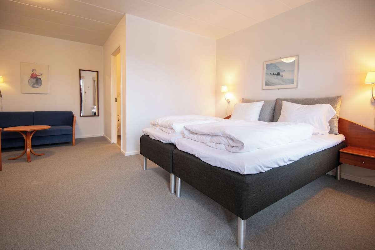 A hotel room with a large bed, a small table, a sofa, and two wall-mounted lamps.