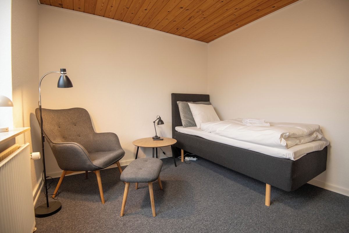 A bedroom with a bed, a chair, a small table, and a floor lamp.