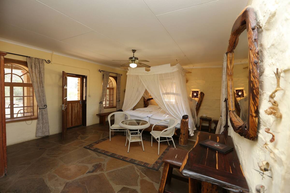 A bedroom with a canopy bed, stone flooring, a round table with chairs, a wooden nightstand, and a large mirror.