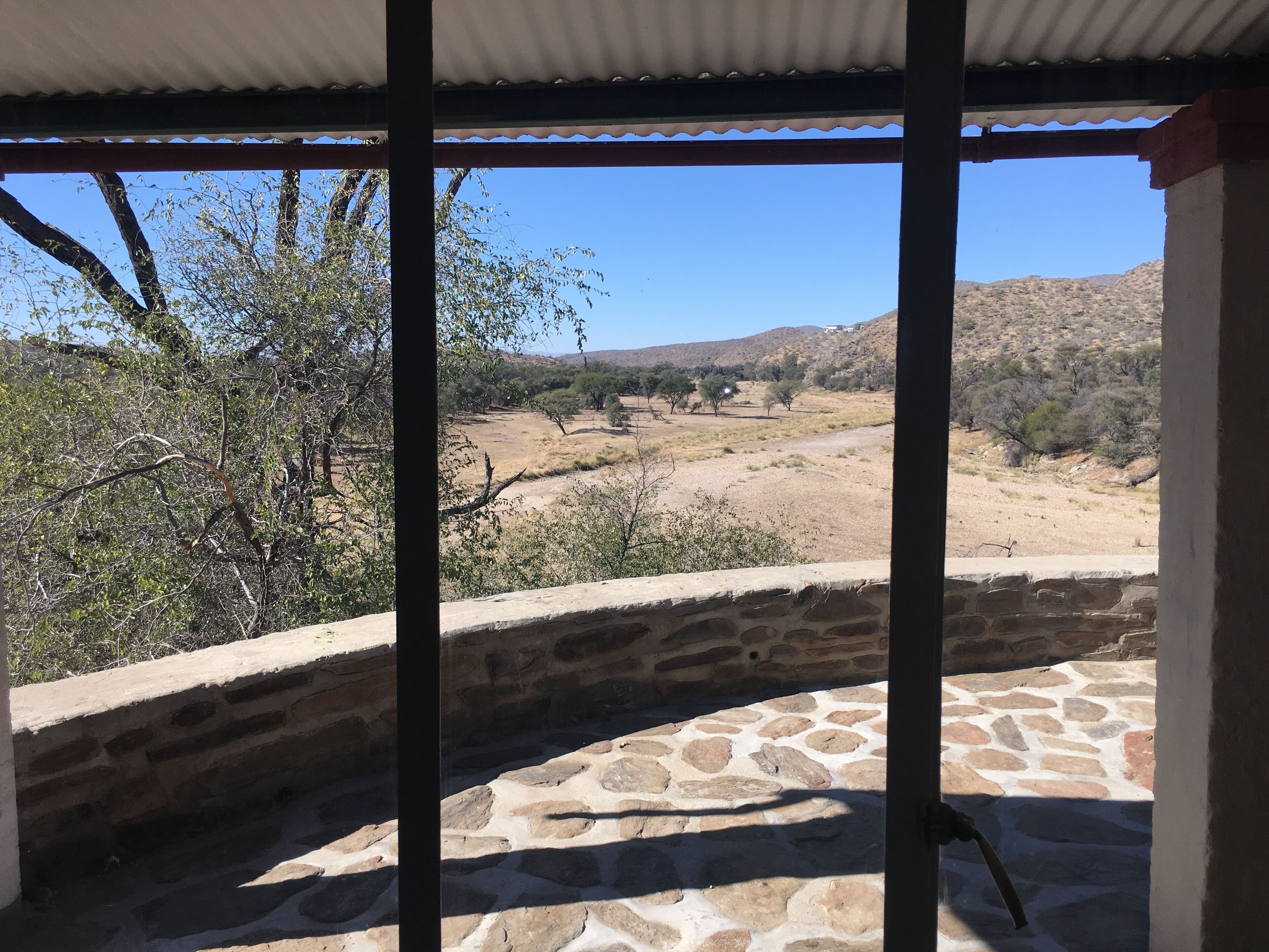 A stone-tiled patio with a view of a dry, hilly landscape through a covered area with metal beams.