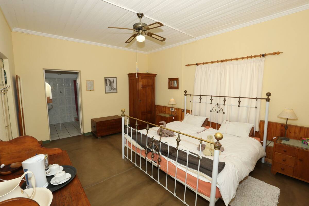 A bedroom with a four-poster bed, wooden furniture, a ceiling fan, and a bathroom visible through an open door.
