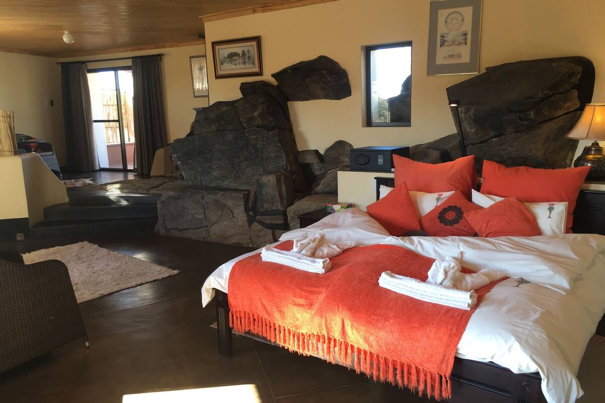 A bedroom with a bed, pillows, a bedside table, a lamp, and a large rock feature.