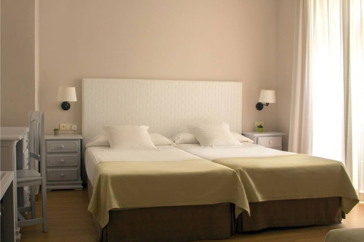 A double bed with a beige headboard, two bedside tables with lamps, a small bedside table with a plant, and a chair.