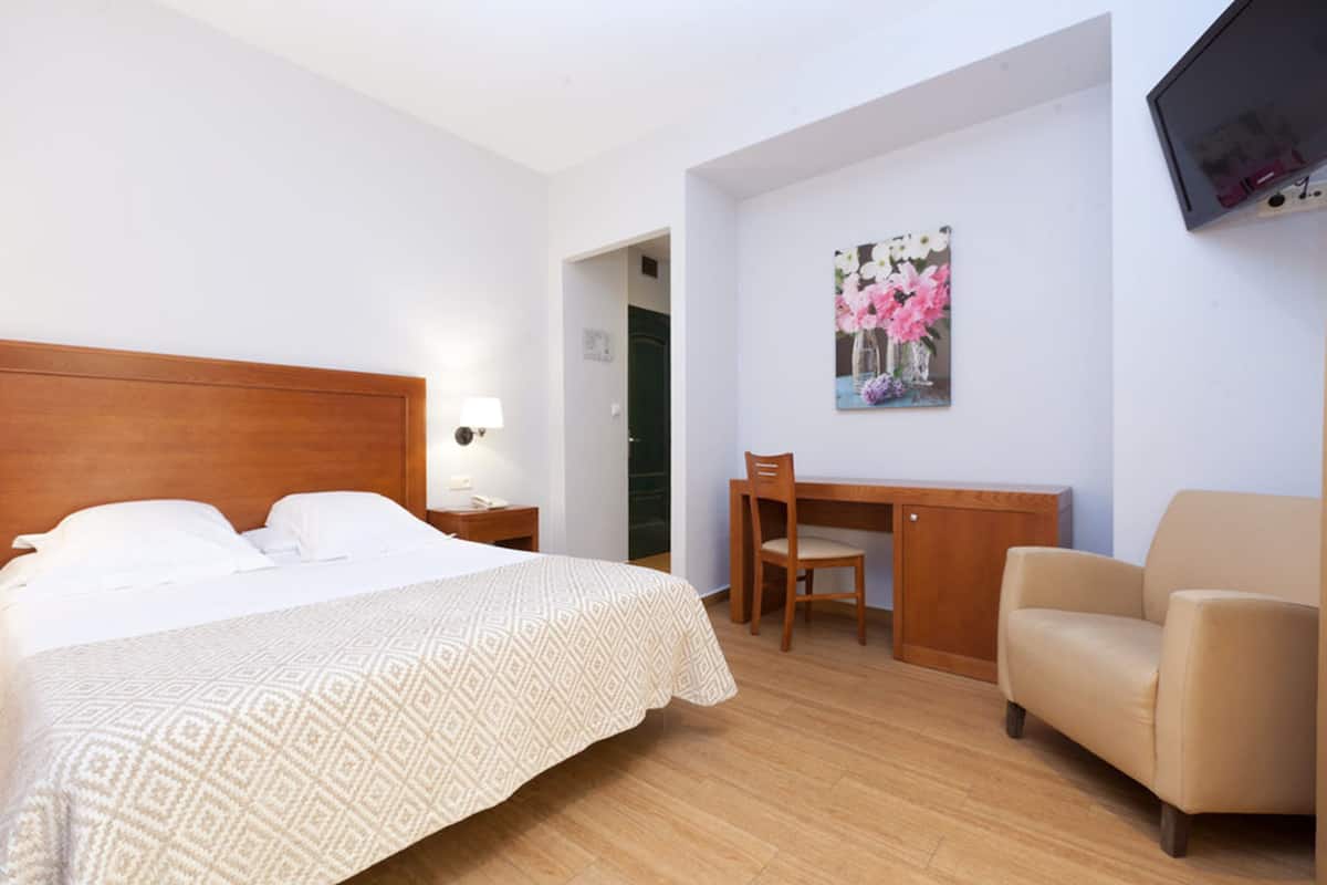A hotel room with a bed, a desk, a chair, and a television on the wall.