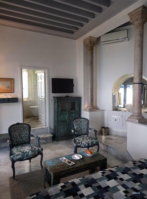 A spacious living room with two patterned armchairs, a wooden coffee table with a bowl of fruit, and a television on the wall.