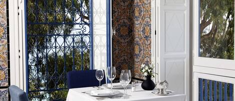 A dining table set with white tablecloth, plates, and wine glasses, with a blue chair and a vase of flowers.