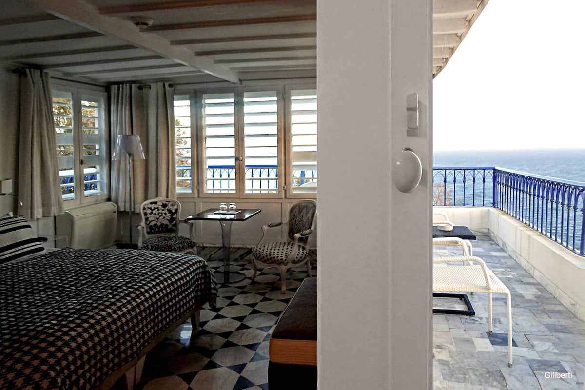 A room with a bed, a table, and two chairs. There is a balcony with a view of the sea.