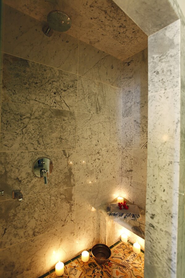 Un baño con paredes de mármol, un cabezal de ducha, un espejo y velas.
