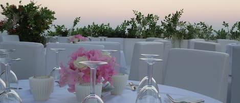 Une table dressée pour un événement formel en plein air, avec des nappes blanches, de la vaisselle transparente et des fleurs roses.