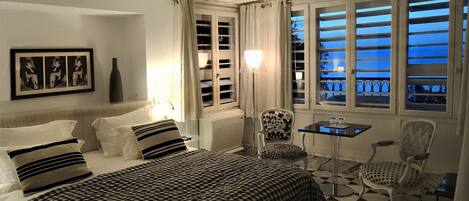A hotel room with a checkered bedspread, a desk with a chair, a chair with a patterned cushion, a framed picture on the wall, and a window with curtains.