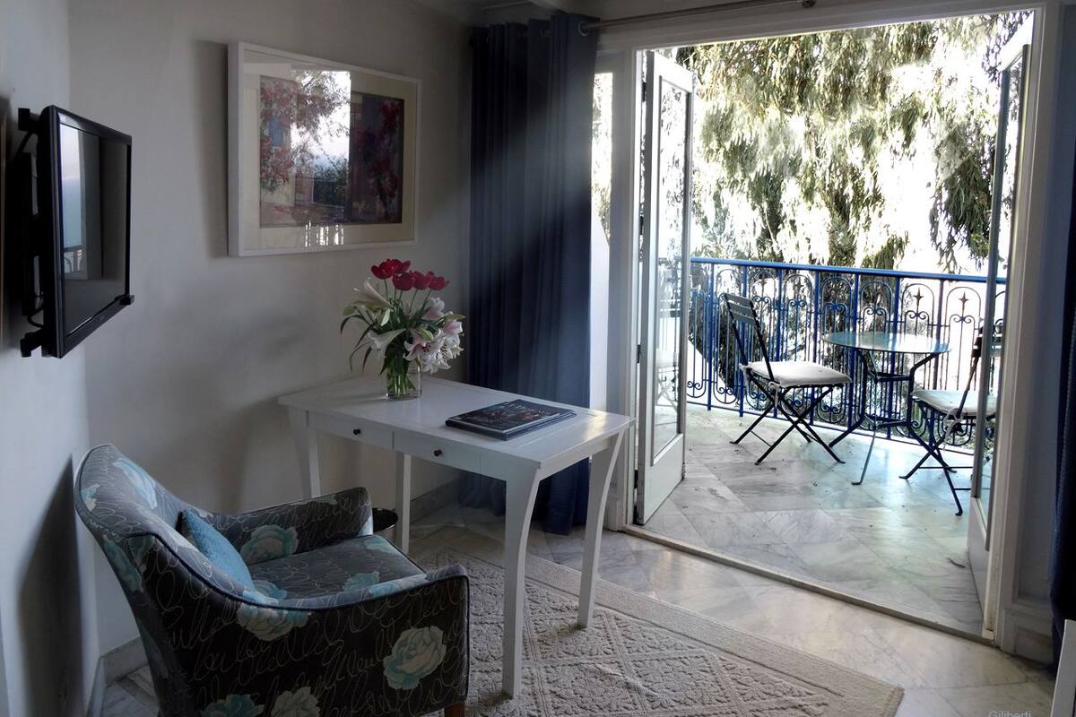 A room with a white desk, a chair, a framed picture, and a balcony with a table and chairs.