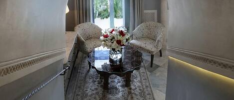 A staircase leading to a room with two patterned armchairs, a glass coffee table with a vase of flowers, and a view through a large window.