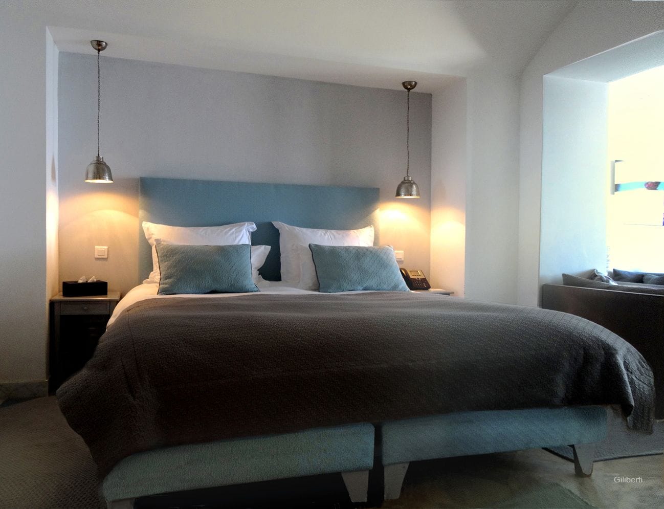Una cama bien hecha con un edredón oscuro y dos almohadas, una mesita de noche con una lámpara, y una ventana con persianas.