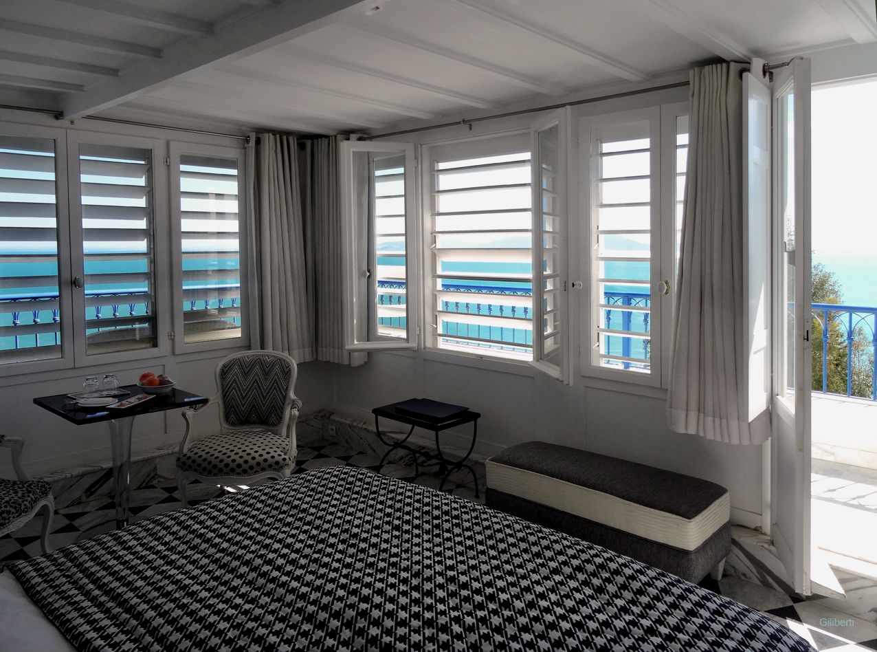 A room with a bed, a chair, a small table, and a bench. There are windows with curtains and a view of the sea.