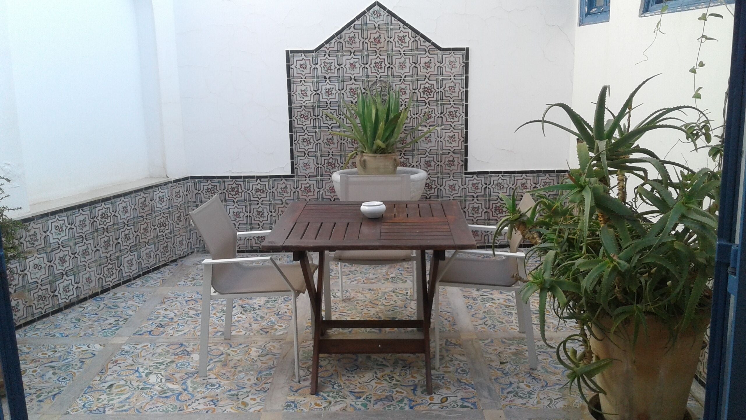 A patio with a wooden table and chairs, a potted plant, and a tiled floor.