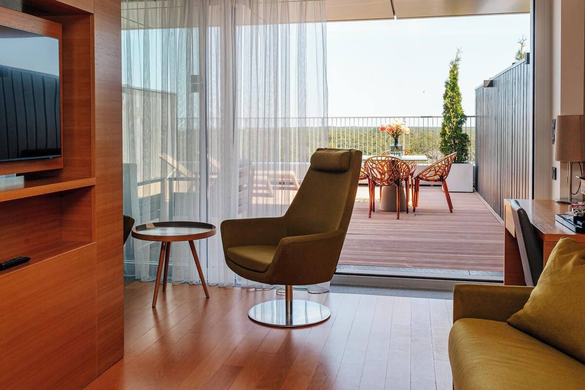 Ein modernes Wohnzimmer mit Fernseher, einer Couch, einem Sessel und einem Tisch mit Stühlen. Es gibt einen Balkon mit Blick auf die Landschaft.