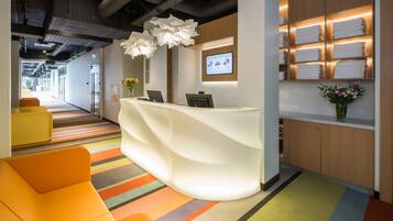 A reception area with a white reception desk, colorful striped carpet, orange seating, and a wall-mounted screen displaying a floral arrangement.