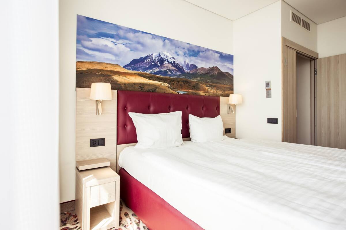 Ein Hotelzimmer mit einem großen Bett, einem roten Kopfteil und einem Wandbild mit einer Berglandschaft.