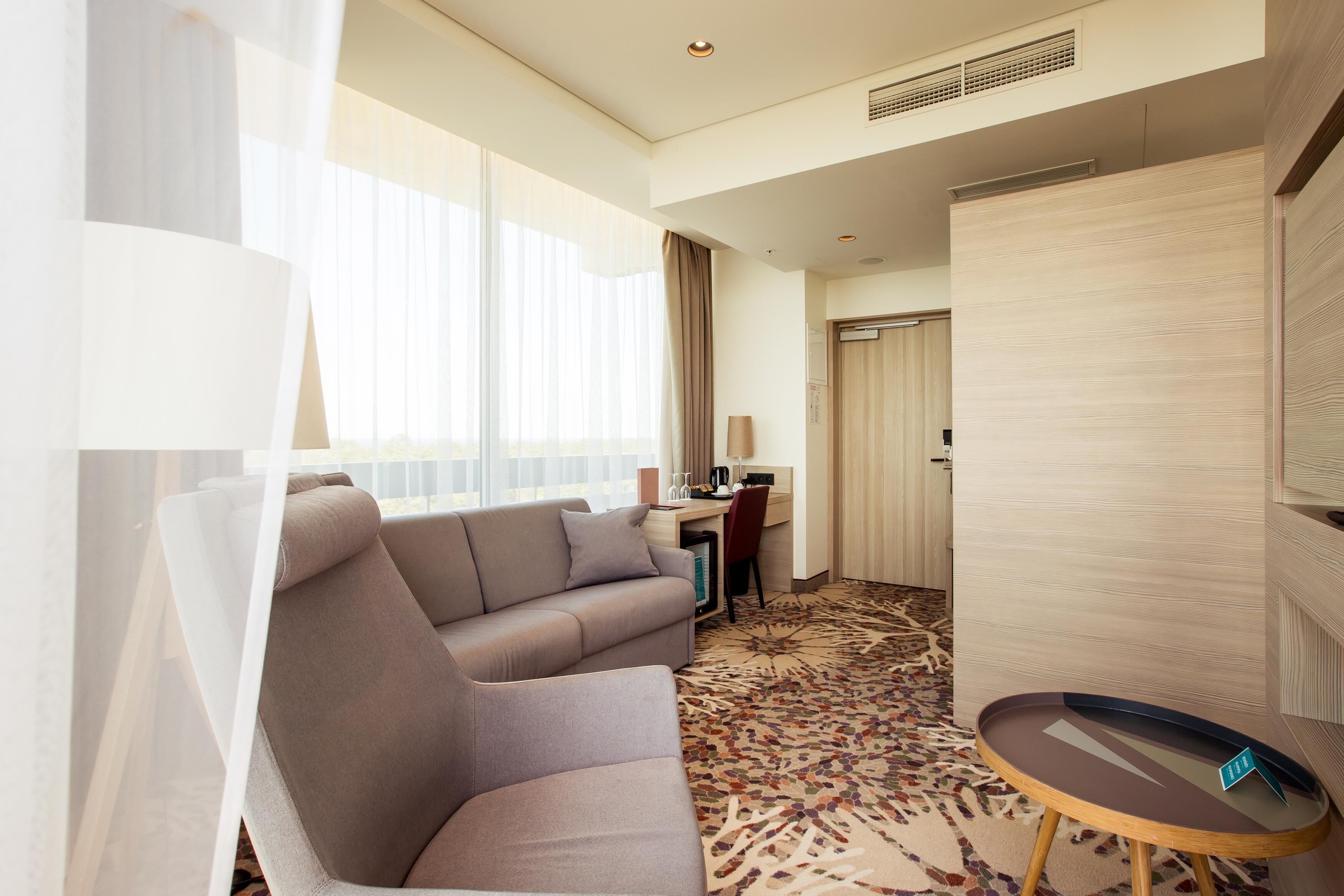 A hotel room with a sofa, a desk, a chair, a small table, and a patterned carpet.