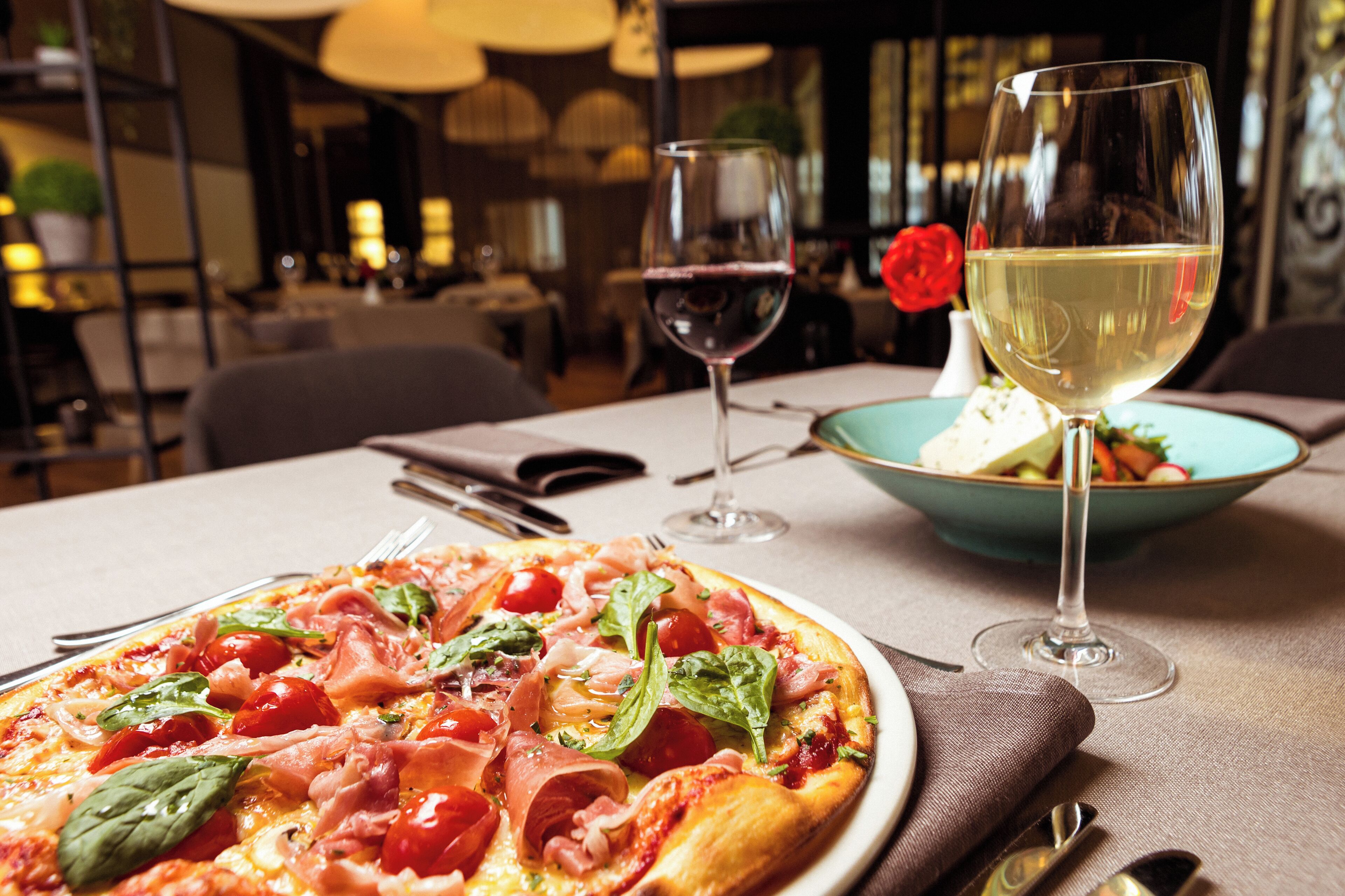 A pizza with various toppings, a glass of red wine, and a bowl of salad on a table in a restaurant setting.