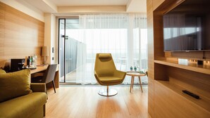 Habitación de hotel moderna con piso de madera, sofá verde, sillón amarillo, una mesita con un control remoto y un televisor sobre un mueble de madera.