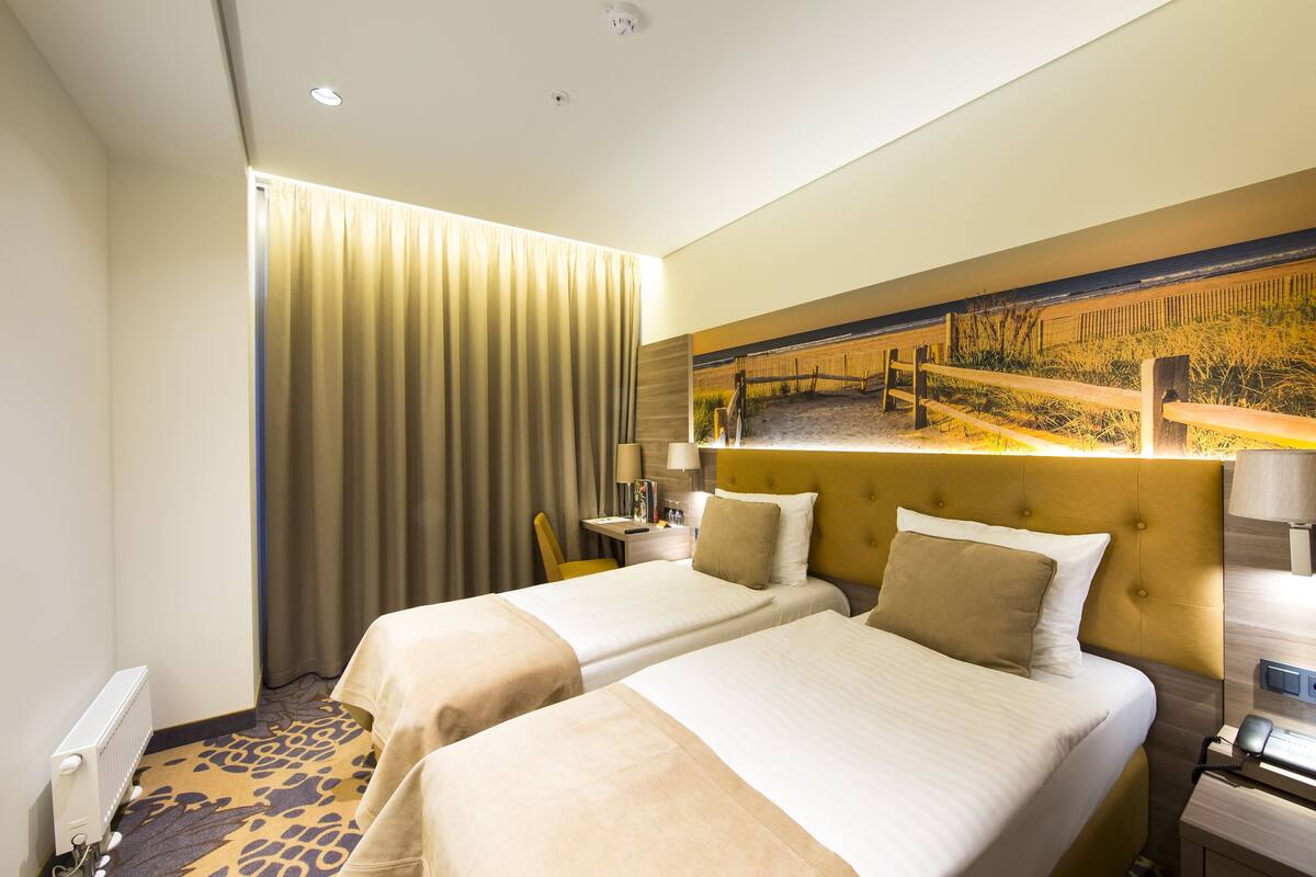 Ein Hotelzimmer mit zwei Betten, einem großen Fenster mit Vorhängen, einem Bettkopfteil mit einem Gemälde und einem Nachttisch mit einer Lampe.