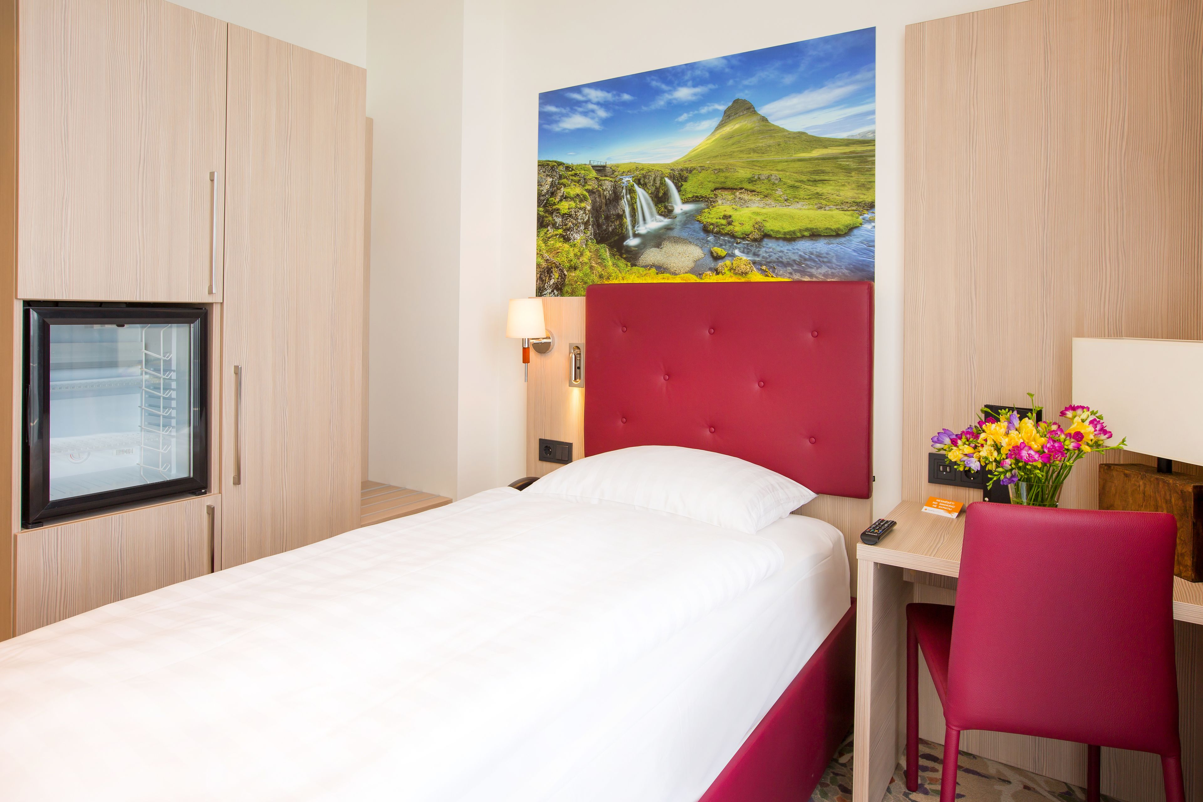 A hotel room with a bed, a desk with a chair, a TV, and a painting of a landscape.