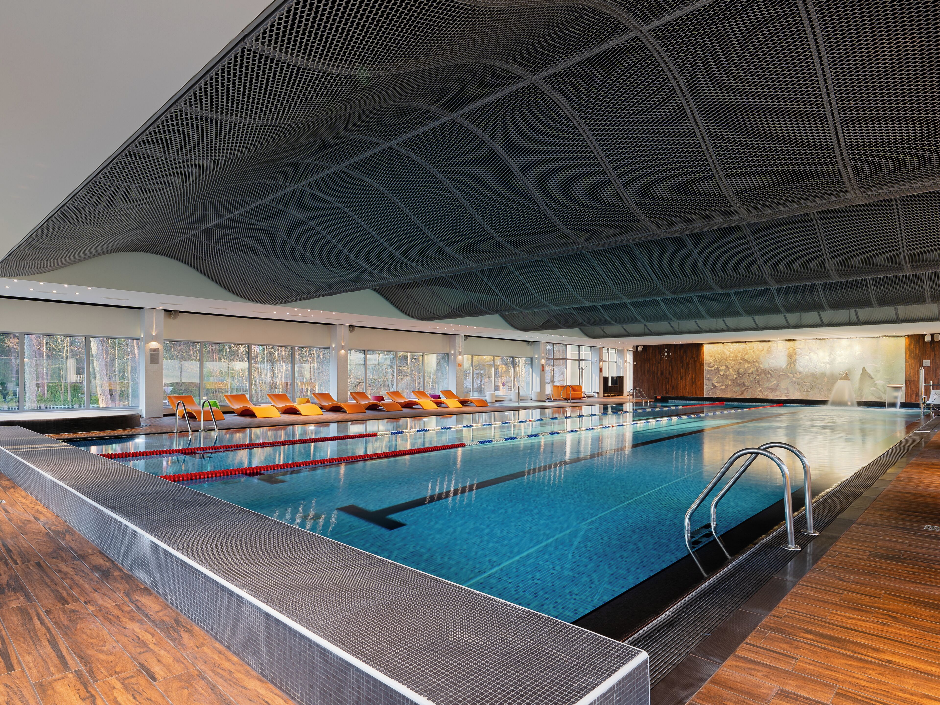 A swimming pool with multiple lanes, lounge chairs, and a view of trees through large windows.