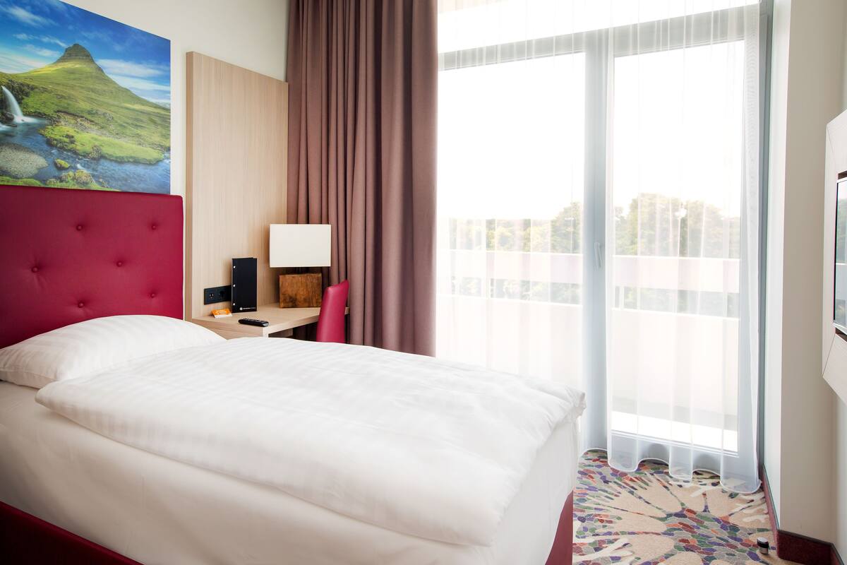 Ein Hotelzimmer mit einem großen Bett, einem Schreibtisch mit Lampe, einem roten Sessel und Blick auf eine Stadtlandschaft durch ein großes Fenster.