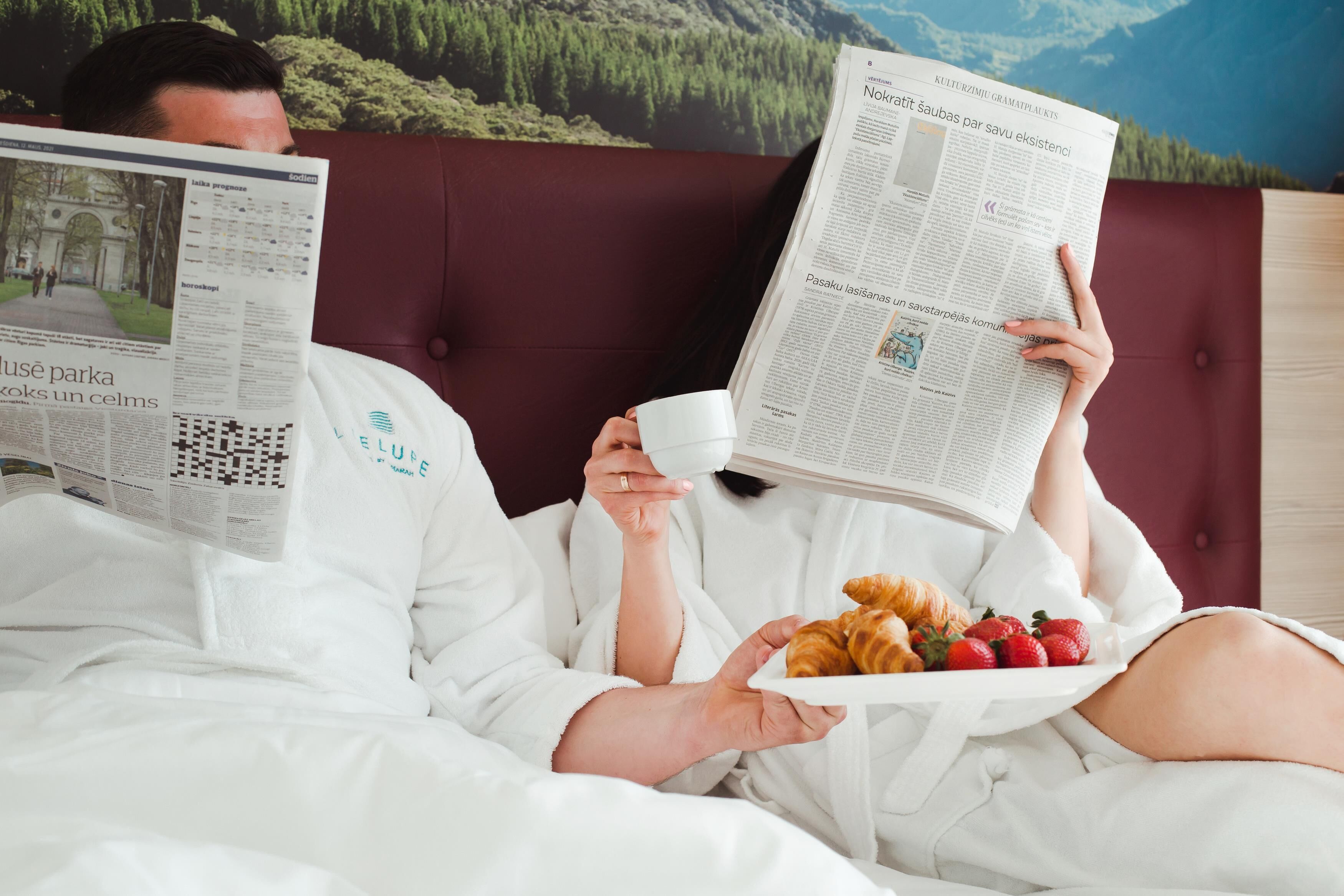 Two people reading newspapers in bed with a scenic view of a forest and mountains in the background.