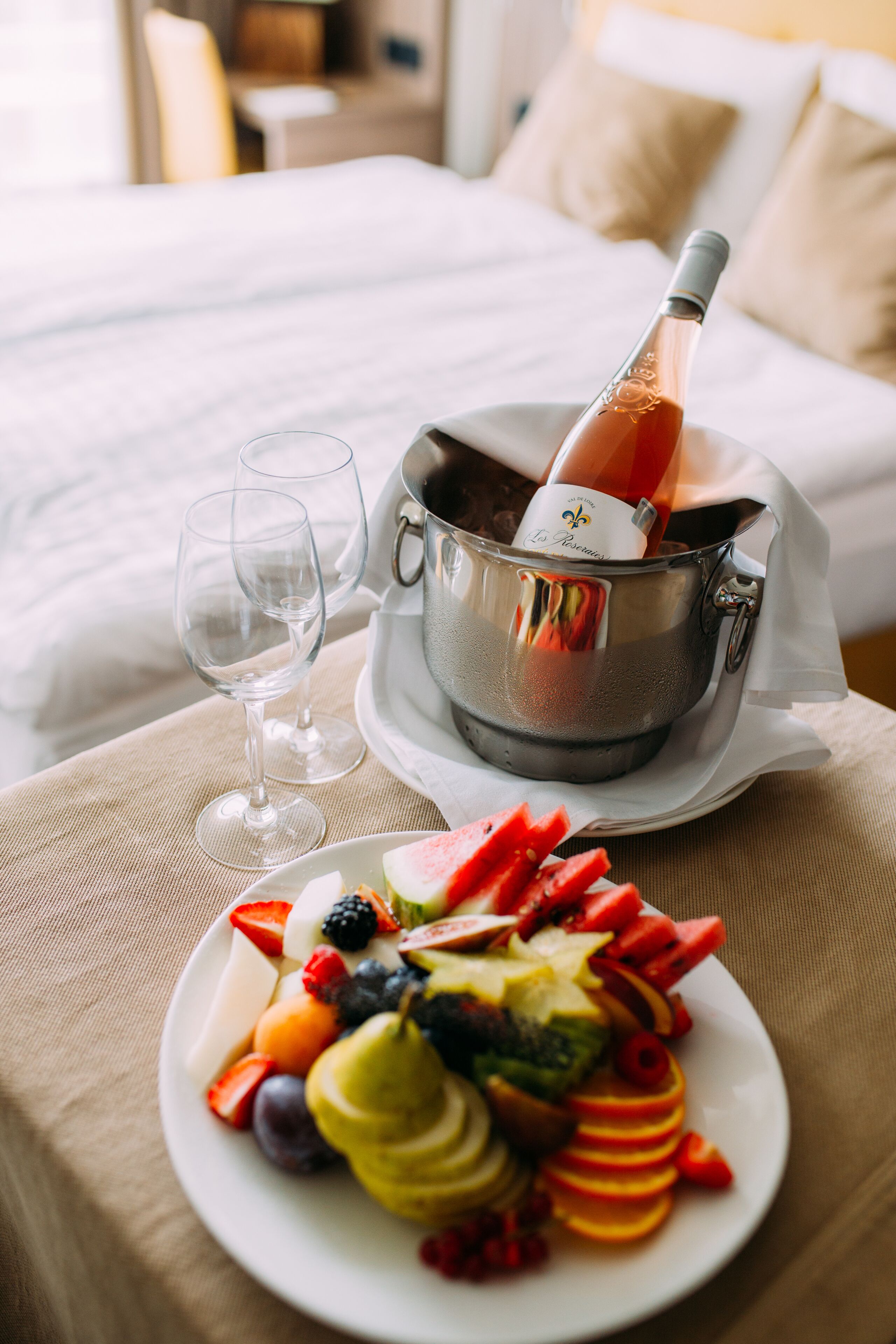A bed with white linens, a bedside table with a bottle of champagne, two glasses, and a plate of fruit.