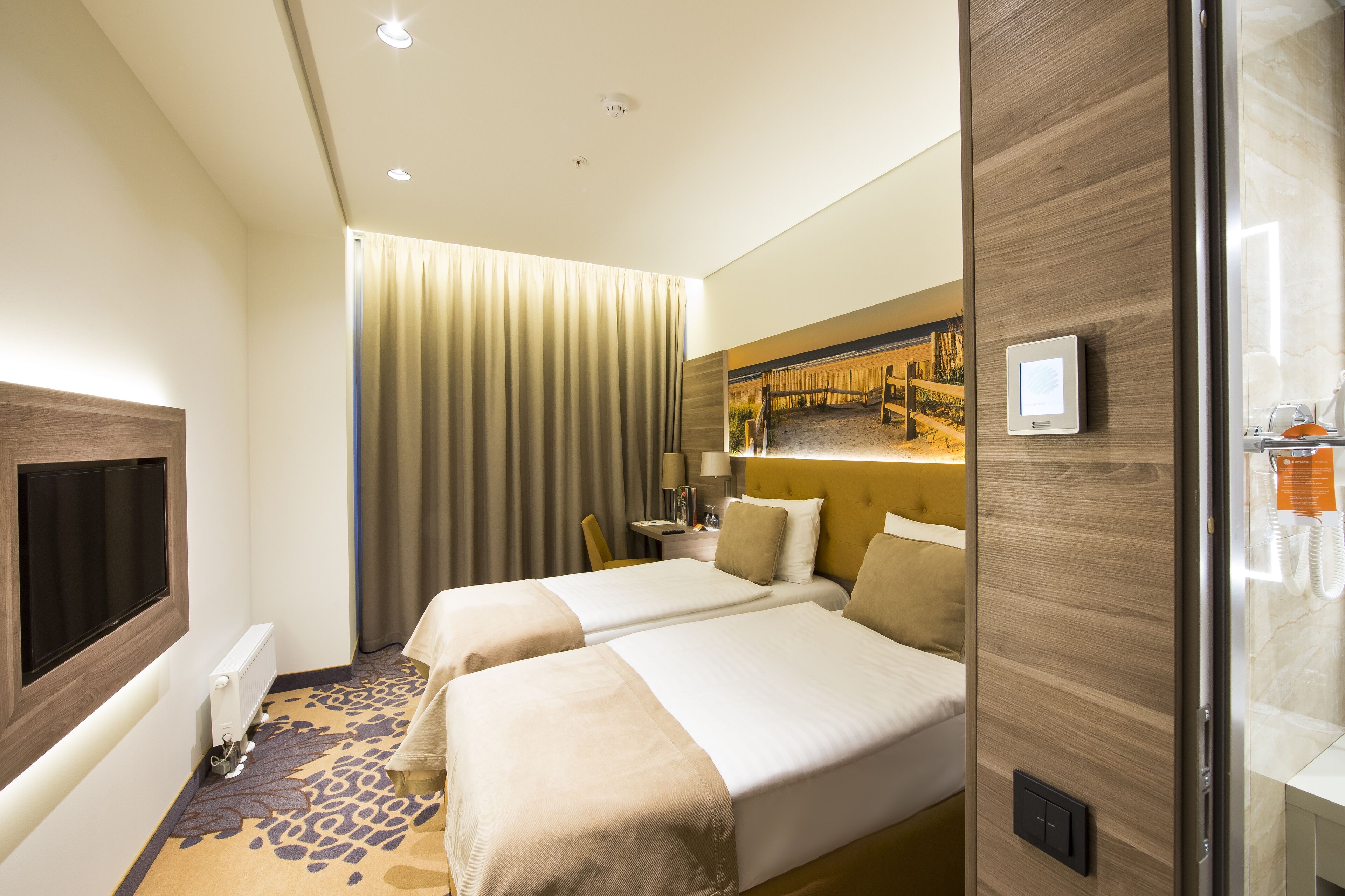 Ein Hotelzimmer mit zwei Betten, einem großen Fenster mit Vorhängen, einem Flachbildfernseher und einer Wand mit Holzvertäfelung.