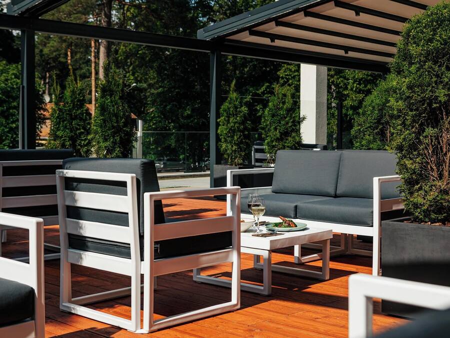Un'area patio esterna con arredi moderni, tra cui sedie e un divano, un tavolo da pranzo con un bicchiere di vino e vista sul verde circostante.