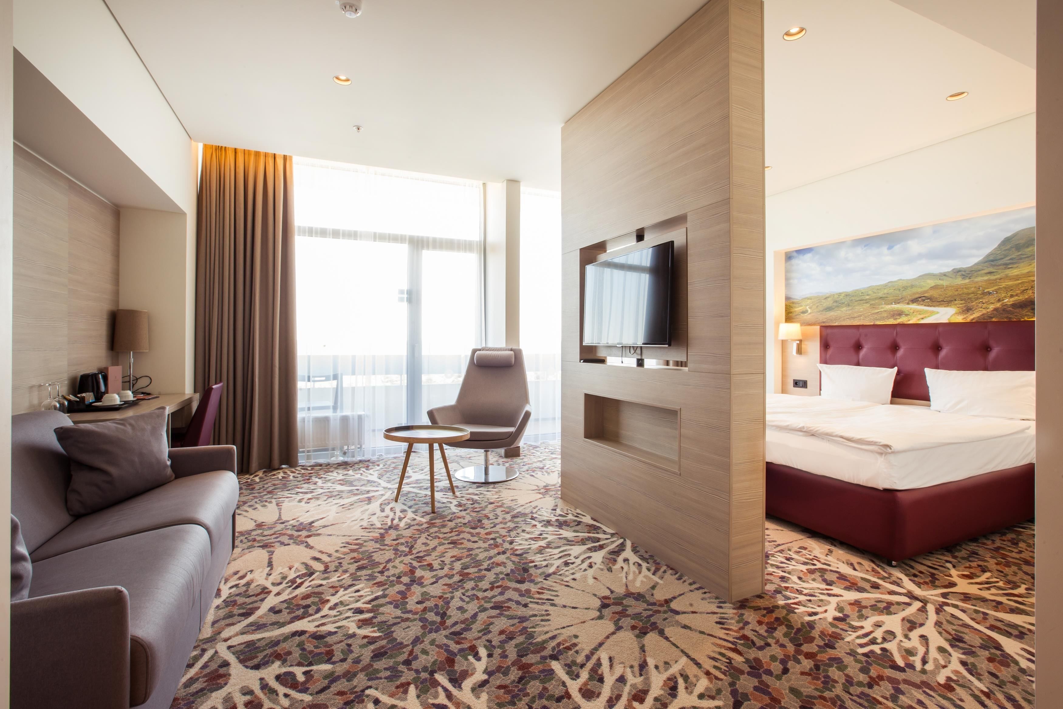 Una habitación de hotel con sofá, sillón, televisión y una cama con cabecera roja.