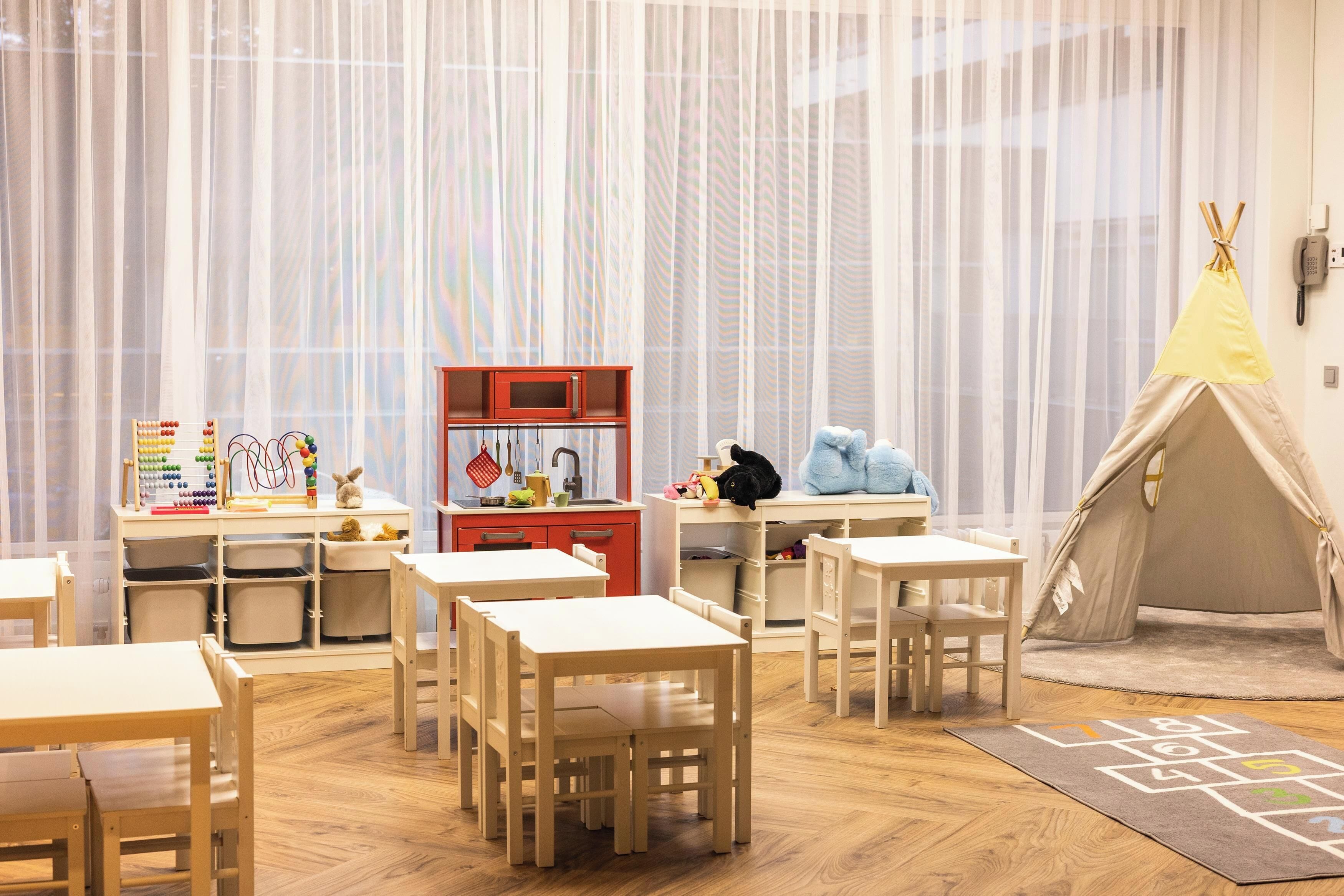 A children's playroom with tables, chairs, a play kitchen, and a teepee.