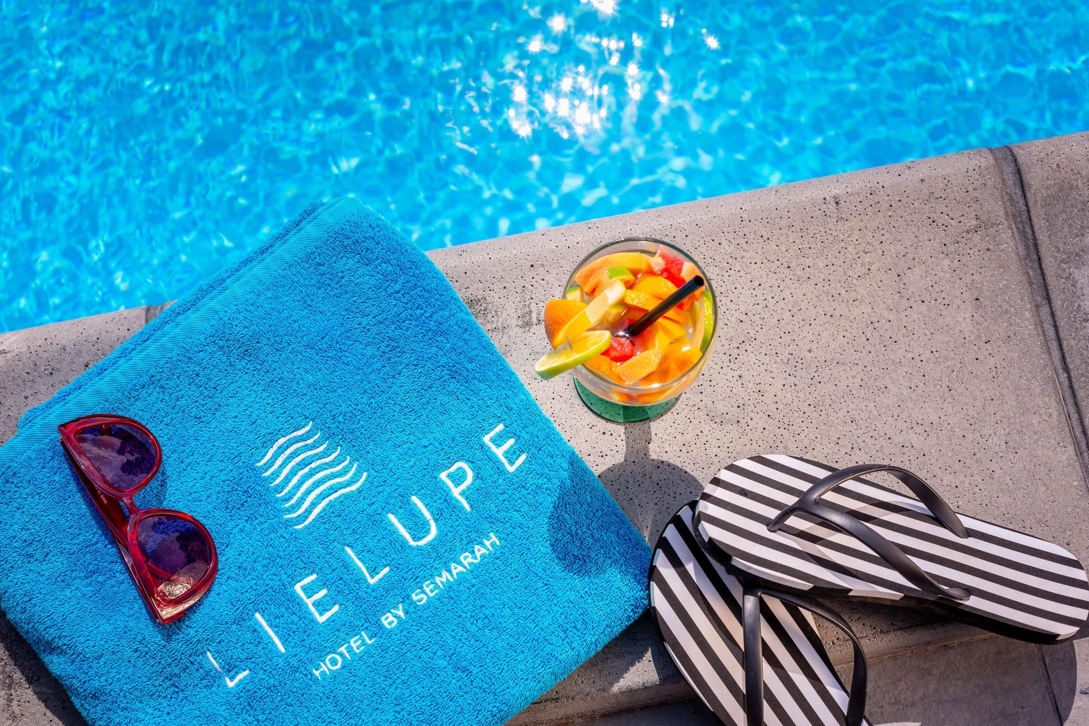 A blue towel with sunglasses and flip-flops on a poolside, and a drink with fruit.