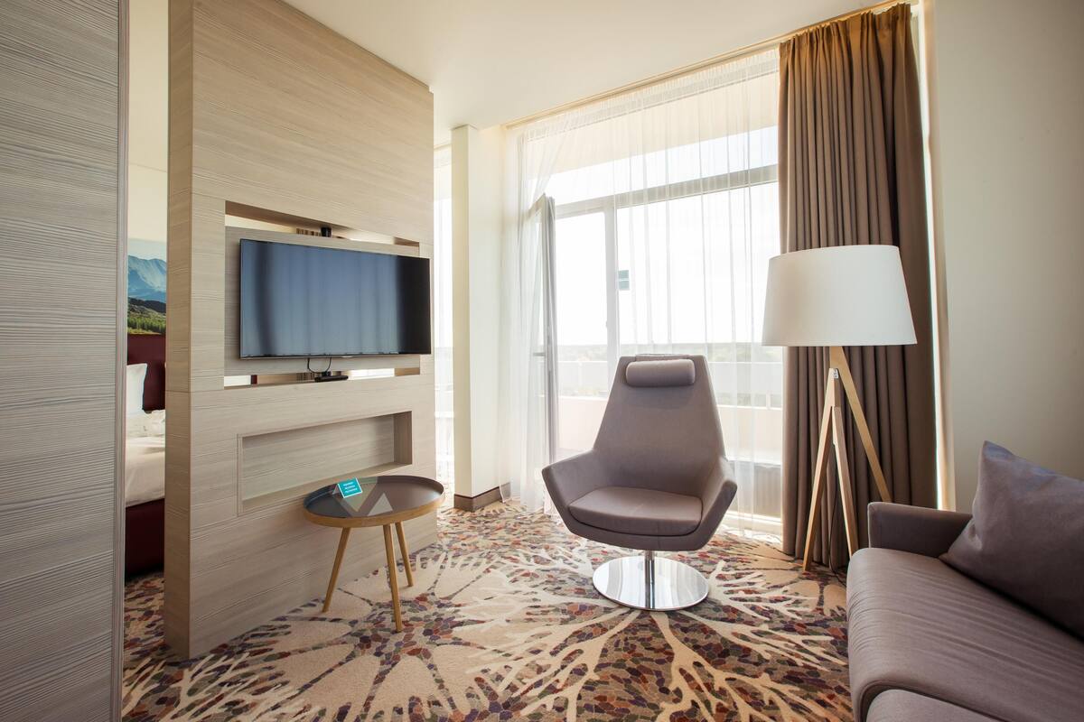 Ein Hotelzimmer mit flachbildfernseher an der Wand, einem modernen Sessel, einer Couch, einer Stehlampe und einem großen Fenster mit Vorhängen.