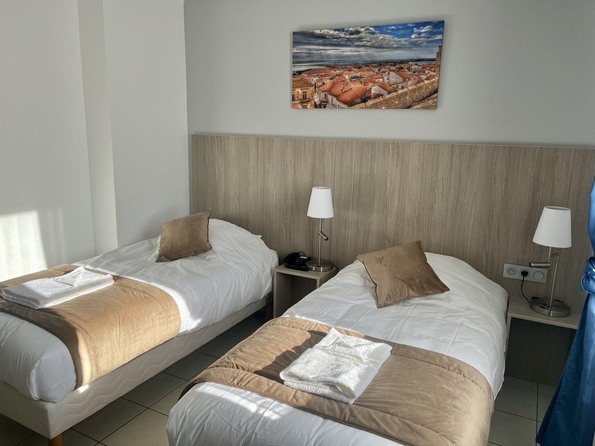 Camera d'albergo con due letti, ciascuno con cuscino e coperta, un comodino con una lampada e un quadro appeso al muro raffigurante una cittadina costiera.
