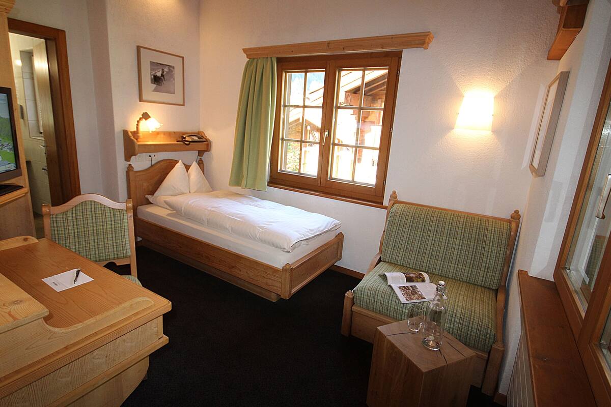 A small, cozy room with a single bed, a wooden desk, a chair, and a window with curtains.