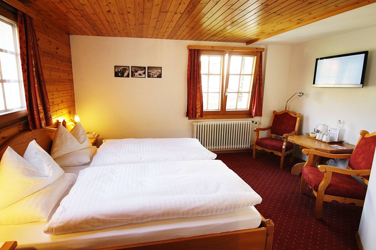 A hotel room with a wooden ceiling, two beds with white linens, a wooden chair, a small table with a coffee maker, and a television on the wall.