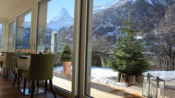 A modern interior with large windows offering a view of snow-covered mountains and a wooden deck outside. - Hotel Welschen Zermatt (Zermatt)